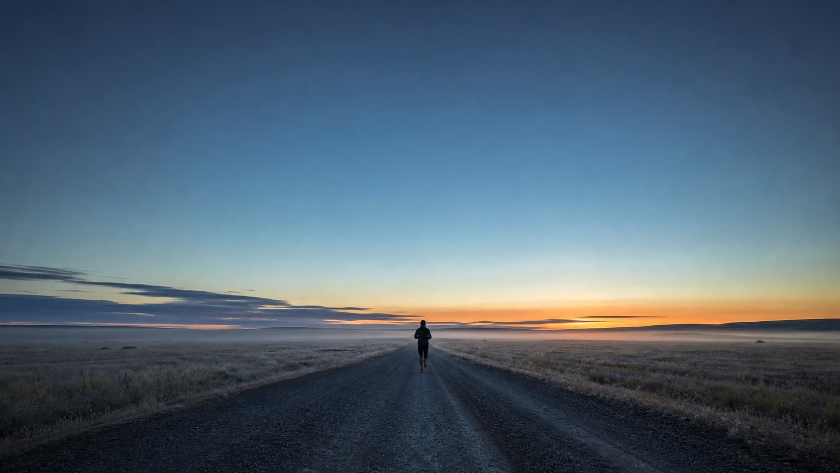 A lone runner heading out along an empty gravel road at dawn, low mist over the open prairie, a warm band of light on the horizon.